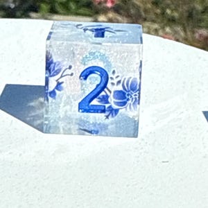 May include: A clear, square die with blue floral designs and the number 2 in blue. The die is made of a translucent material, allowing light to pass through. The floral patterns are reminiscent of porcelain designs.