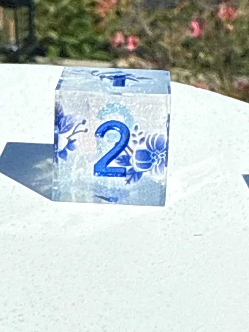 May include: A clear, square die with blue floral designs and the number 2 in blue. The die is made of a translucent material, allowing light to pass through. The floral patterns are reminiscent of porcelain designs.