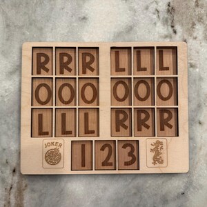 May include: A rectangular light-colored wooden game board featuring a grid of eighteen squares with the letters R, O, and L engraved in dark brown. Below the grid are three additional squares labeled 1, 2, and 3, flanked by a Joker icon and a dragon icon.
