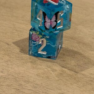 May include: Two stacked, clear blue resin dice with silver numbering and floral and butterfly inclusions. The top die features a butterfly and the numbers 4 and 5. The bottom die shows the number 2 and a pink rose.