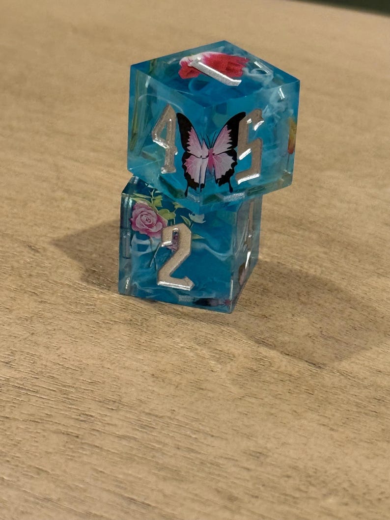 May include: Two stacked, clear blue resin dice with silver numbering and floral and butterfly inclusions. The top die features a butterfly and the numbers 4 and 5. The bottom die shows the number 2 and a pink rose.