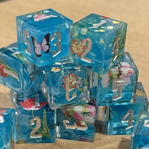 May include: A collection of clear, blue resin dice with silver numbers and floral and butterfly inclusions. The dice are cube-shaped and feature various flowers and a butterfly. The numbers are in a silver, serif font.