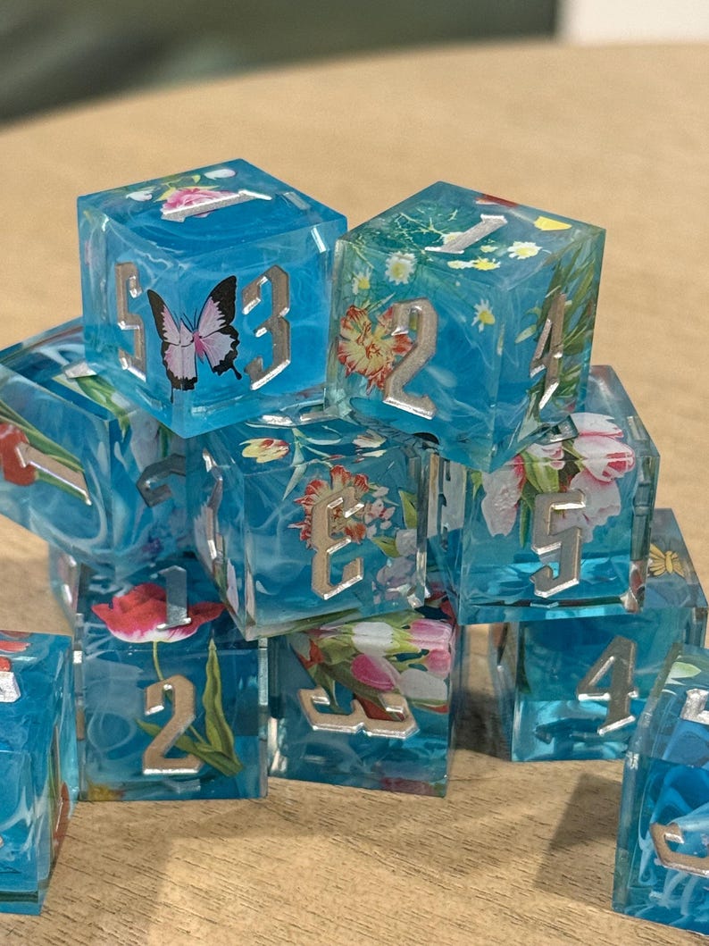 May include: A collection of clear, blue resin dice with silver numbers and floral and butterfly inclusions. The dice are cube-shaped and feature various flowers and a butterfly. The numbers are in a silver, serif font.