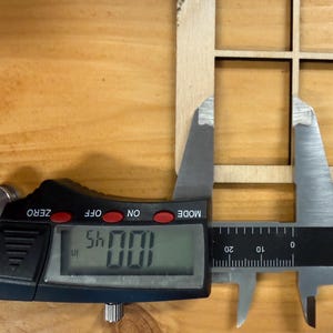 May include: A light-colored wooden grid panel with square cutouts being measured by a black and silver digital caliper. The digital display clearly shows a measurement of 1.0045 inches, indicating the precise width of the wooden frame section.