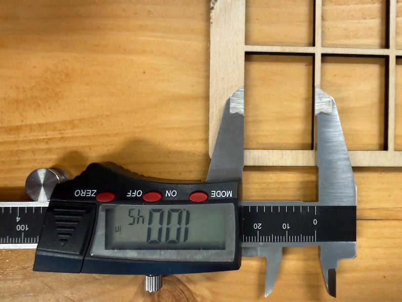 May include: A light-colored wooden grid panel with square cutouts being measured by a black and silver digital caliper. The digital display clearly shows a measurement of 1.0045 inches, indicating the precise width of the wooden frame section.