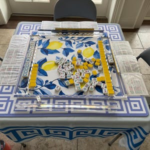 May include: A Mahjong set on a table with a blue and white patterned tablecloth. The game board features a lemon and leaf design. Yellow and white tiles are scattered across the board, with yellow tile racks and score sheets.