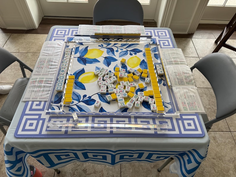 May include: A Mahjong set on a table with a blue and white patterned tablecloth. The game board features a lemon and leaf design. Yellow and white tiles are scattered across the board, with yellow tile racks and score sheets.