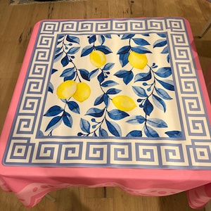 May include: A square tablecloth with a pink border and a white center featuring a lemon and leaf design. The center design has yellow lemons and blue leaves. The border has a Greek key pattern in blue.