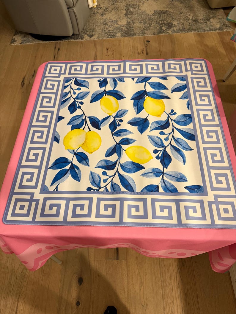 May include: A square tablecloth with a pink border and a white center featuring a lemon and leaf design. The center design has yellow lemons and blue leaves. The border has a Greek key pattern in blue.