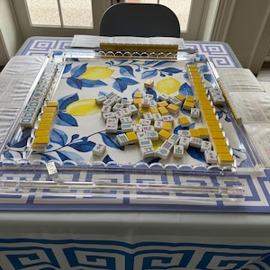 May include: A Mahjong set on a table with a blue and white lemon pattern. The set includes yellow tiles, clear acrylic racks, and a dice. The table is covered with a blue and white geometric patterned tablecloth.