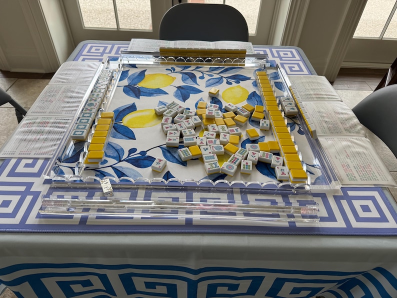 May include: A Mahjong set on a table with a blue and white lemon pattern. The set includes yellow tiles, clear acrylic racks, and a dice. The table is covered with a blue and white geometric patterned tablecloth.