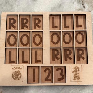 May include: A rectangular light-colored wooden board featuring a grid of engraved letters and numbers. The top section contains three rows of letters R, O, and L. The bottom section displays a Joker icon, the numbers 1, 2, 3, and a dragon icon.