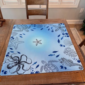May include: A square, light blue mat with an ocean theme featuring illustrations of sea creatures like starfish, turtles, and an octopus. The mat has a central starfish design and is surrounded by various marine life. The website address is visible in the bottom right corner.