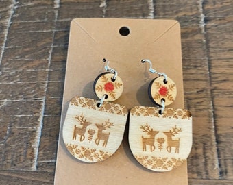 May include: Wooden earrings with a festive design. Each earring features a circular top with a red floral accent and a larger, curved bottom piece. The bottom piece has a laser-cut design of two reindeer and floral patterns.