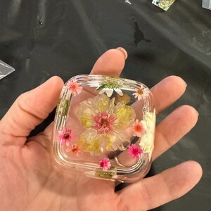 May include: A clear, square resin object with embedded dried flowers in shades of pink, yellow, and white. The flowers are arranged inside the transparent resin, creating a decorative and artistic design. Additional resin objects with flowers are visible in the background.
