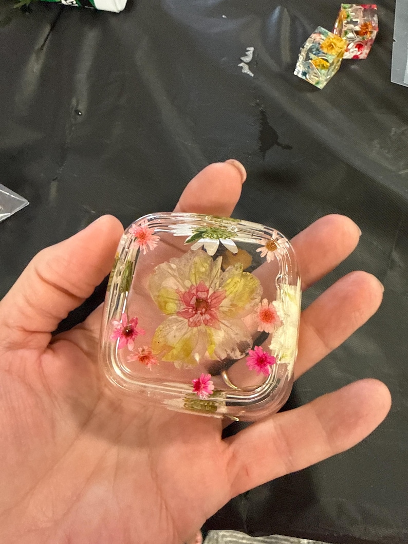 May include: A clear, square resin object with embedded dried flowers in shades of pink, yellow, and white. The flowers are arranged inside the transparent resin, creating a decorative and artistic design. Additional resin objects with flowers are visible in the background.