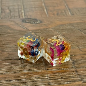 May include: Two clear resin dice with gold numbers and colorful dried flowers embedded inside. The dice are hexagonal and sit on a wooden surface. One die shows the number 6, the other shows the number 2.