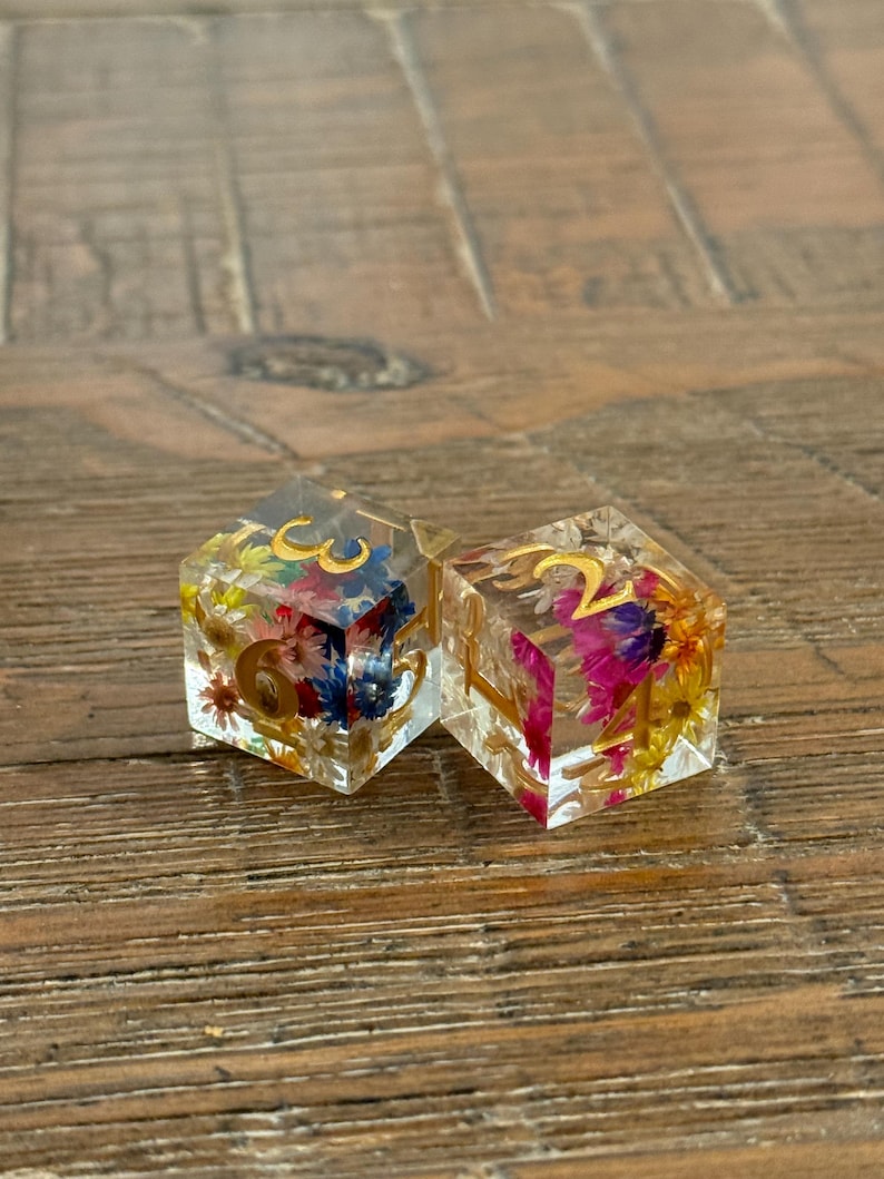May include: Two clear resin dice with gold numbers and colorful dried flowers embedded inside. The dice are hexagonal and sit on a wooden surface. One die shows the number 6, the other shows the number 2.