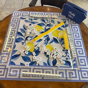 May include: A Mahjong set with a blue and white floral design. The set includes a blue carrying case with "Mahj Tiles" printed on it, yellow tile racks, and numerous tiles scattered on the playing surface. The board has a blue and white geometric border.