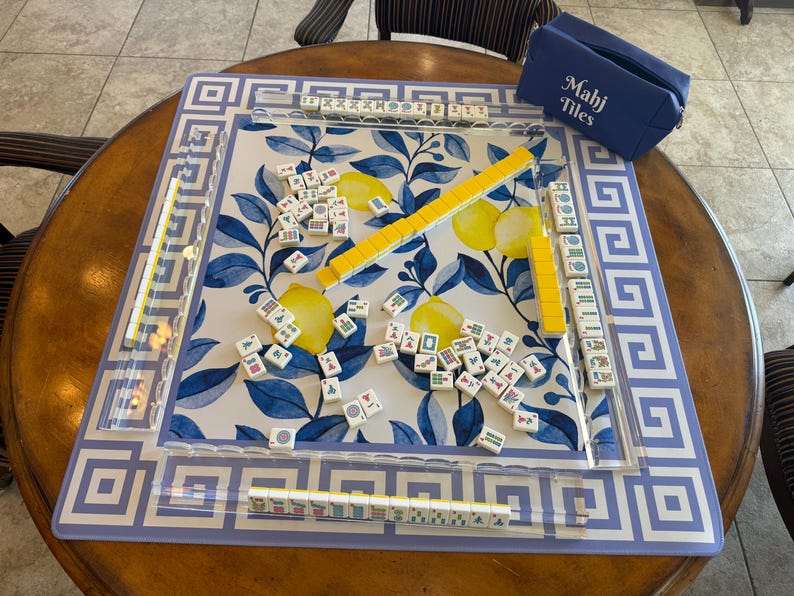 May include: A Mahjong set with a blue and white floral design. The set includes a blue carrying case with "Mahj Tiles" printed on it, yellow tile racks, and numerous tiles scattered on the playing surface. The board has a blue and white geometric border.