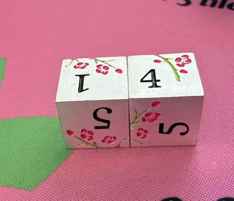 May include: Two white wooden dice with black numbers and pink floral accents. One die shows the numbers 1 and 5, the other shows 4 and 5. The dice are on a pink surface.