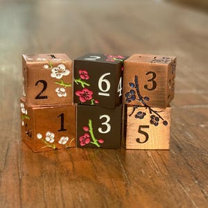 May include: Six copper-colored dice with white and pink floral designs. Each die has a different number from 1 to 6. The dice are arranged on a wooden surface.