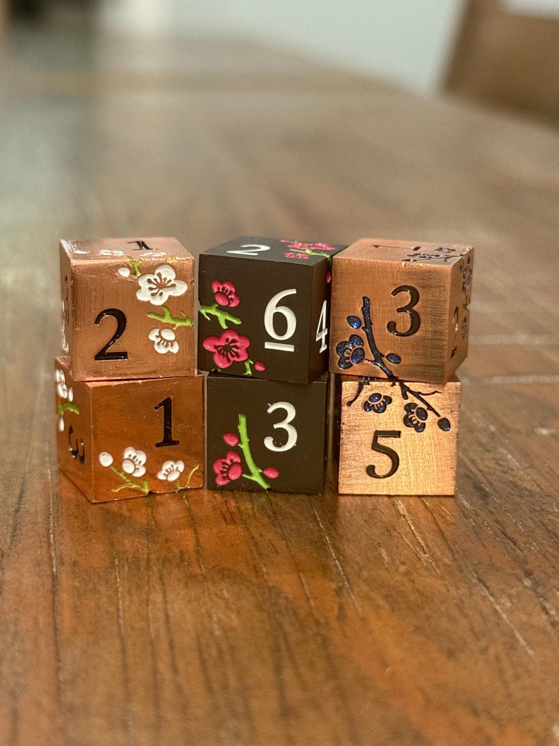 May include: Six copper-colored dice with white and pink floral designs. Each die has a different number from 1 to 6. The dice are arranged on a wooden surface.