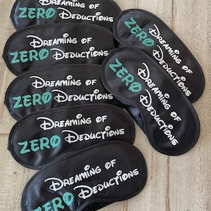 May include: Six black sleep masks with white text that reads "Dreaming of Zero Deductions" in a whimsical font. The text is outlined in teal glitter.