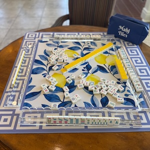 May include: Mahjong game set on a round wooden table. The game board features a blue and white floral design with a Greek key border. The set includes tiles, yellow tile racks, and a blue carrying case with "Mahj Tiles" printed on it.