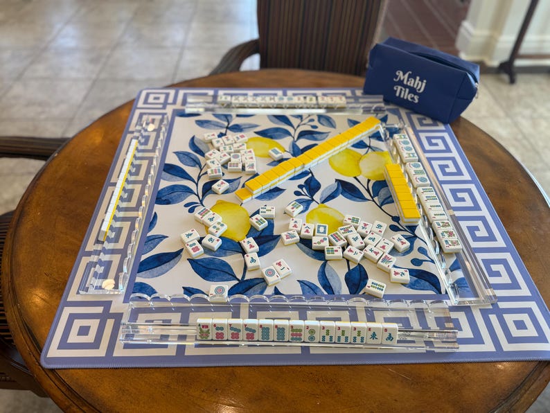 May include: Mahjong game set on a round wooden table. The game board features a blue and white floral design with a Greek key border. The set includes tiles, yellow tile racks, and a blue carrying case with "Mahj Tiles" printed on it.