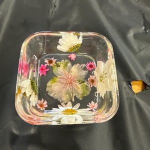 May include: Clear square resin dish with embedded dried flowers in shades of pink, white, and green. The flowers are visible through the transparent resin, creating a decorative and artistic piece. The dish has rounded corners and a slightly raised edge.