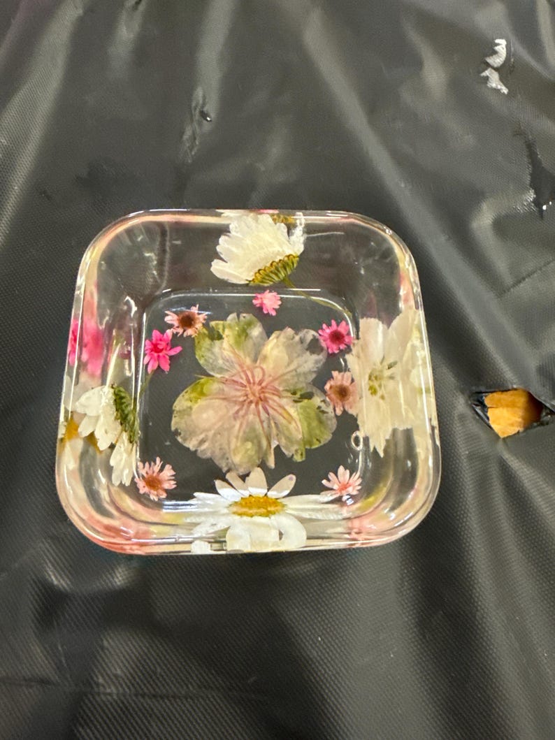 May include: Clear square resin dish with embedded dried flowers in shades of pink, white, and green. The flowers are visible through the transparent resin, creating a decorative and artistic piece. The dish has rounded corners and a slightly raised edge.