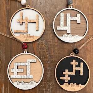 Op de afbeelding: Vier houten kerstversieringen met gepersonaliseerde namen in een Scrabble-achtig ontwerp. Elke versiering heeft een andere achtergrondkleur: wit, bruin, lichtbruin en zwart. De versieringen zijn rond met een houten frame en een touwtje om op te hangen.