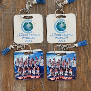 May include: Two white square keychains with blue tassels. Each keychain has a logo for "The Cheerleading Worlds 2023" with a globe graphic. The keychains also feature a photo of a cheerleading squad in blue and white uniforms.