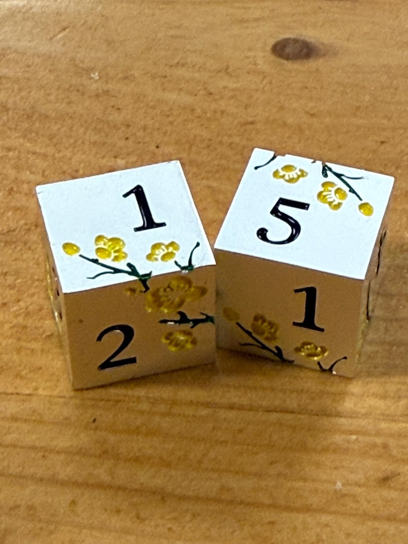 May include: Two white dice with black numbers and yellow floral accents. One die shows the numbers 1, 2, and 1. The other die shows the numbers 5, 1, and 1. The dice are on a wooden surface.