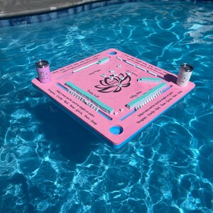 Puede incluir: Tablero de juego flotante rosa con un dise&ntilde;o de flor de loto y fichas azules y verdes. El tablero tiene instrucciones para jugar al juego, "Los comodines no se pueden usar en individuales o en pares. Dragones:  Crazy 8, Rojo, Azul, Verde, Rosa, Salvia."