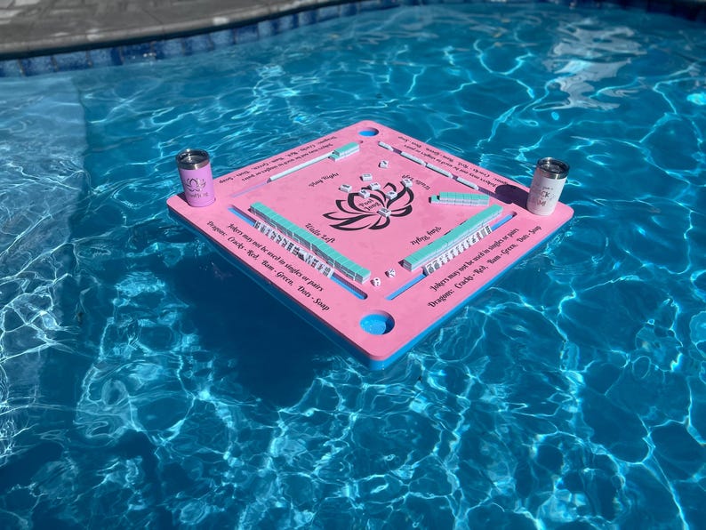 Puede incluir: Tablero de juego flotante rosa con un dise&ntilde;o de flor de loto y fichas azules y verdes. El tablero tiene instrucciones para jugar al juego, "Los comodines no se pueden usar en individuales o en pares. Dragones:  Crazy 8, Rojo, Azul, Verde, Rosa, Salvia."