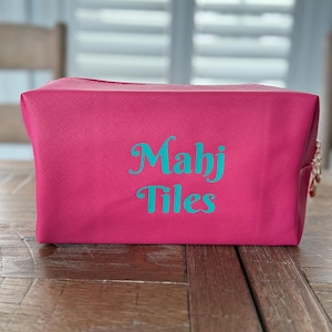 May include: A rectangular, magenta-colored cosmetic bag with the words "Mahj Tiles" in teal script. The bag has a textured surface and a zipper closure with a gold-colored pull. The bag is sitting on a wooden surface.