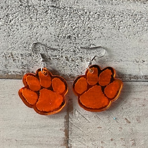 May include: A pair of orange paw print earrings with silver hooks. The paw prints are shiny and have a 3D effect.