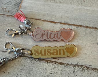 May include: Two acrylic keychains with silver-toned lobster clasps. The top keychain is rose gold with the name Erica and a heart icon. The bottom keychain is gold with the name Susan and a heart icon. The top keychain includes a pink tassel.