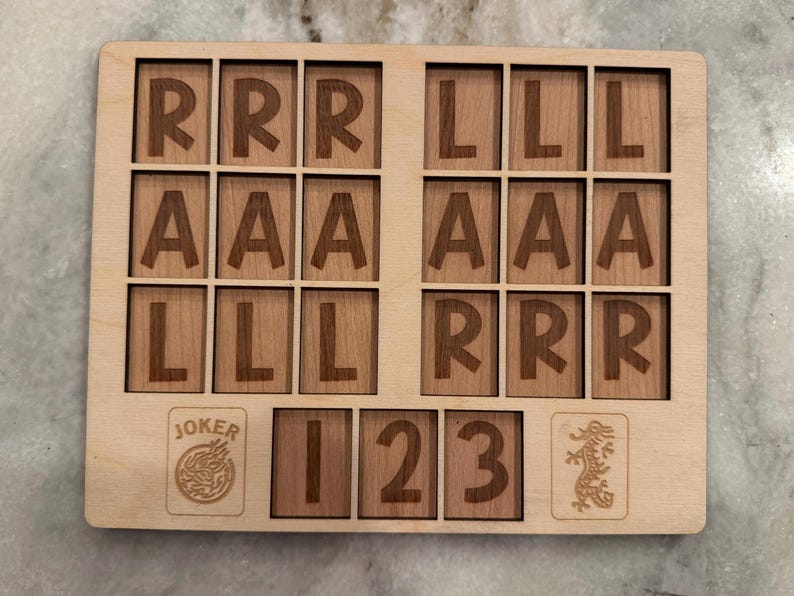 May include: A rectangular light-colored wooden board featuring a grid of laser-engraved letters and numbers. The top section contains three rows of letters R, A, and L. The bottom section displays a Joker icon, the numbers 1, 2, 3, and a dragon icon.