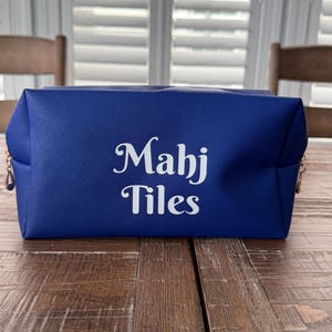 May include: A royal blue rectangular cosmetic bag with the words "Mahj Tiles" in white script. The bag has gold-colored zipper pulls and sits on a wooden table.