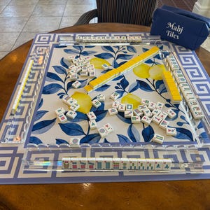 May include: A Mahjong game set on a round wooden table. The game board features a blue and white floral design with a geometric border. Yellow and white tiles are scattered across the board, with a blue carrying case that reads "Mahj Tiles".