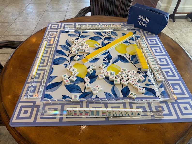 May include: A Mahjong game set on a round wooden table. The game board features a blue and white floral design with a geometric border. Yellow and white tiles are scattered across the board, with a blue carrying case that reads "Mahj Tiles".