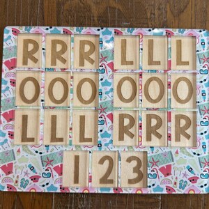 May include: A rectangular wooden puzzle board featuring a colorful summer-themed pattern of flip-flops, sunglasses, and watermelons. The board contains cutouts holding wooden tiles engraved with the letters R, L, O, and the numbers 1, 2, and 3.