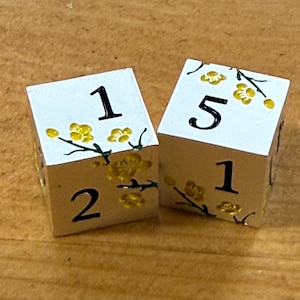 May include: Two white dice with black numbers and yellow floral accents. The dice show the numbers 1, 2, and 5. The dice are decorated with yellow flowers and green stems.