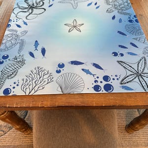 May include: A square, marine-themed placemat featuring a light blue gradient background. The placemat is decorated with black and white illustrations of sea creatures, including an octopus, starfish, sea turtles, fish, and a seahorse. Bubbles and coral complete the design.