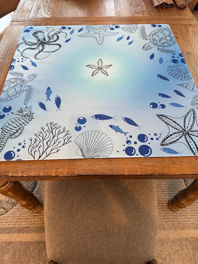 May include: A square, marine-themed placemat featuring a light blue gradient background. The placemat is decorated with black and white illustrations of sea creatures, including an octopus, starfish, sea turtles, fish, and a seahorse. Bubbles and coral complete the design.