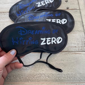 May include: Black sleep masks with the text "Dreaming of Zero" in blue and white. The sleep masks have an elastic band and are designed to block out light. The masks are made of a soft, dark material.