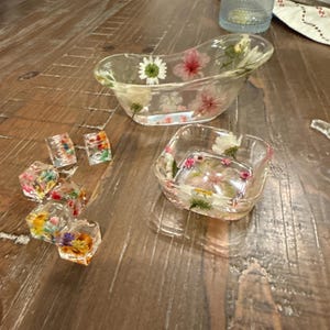May include: Clear resin decorative pieces with embedded dried flowers. The image shows a small bathtub-shaped dish, a square dish, and several small, cube-shaped resin pieces. The flowers are various colors, including white, pink, and yellow, set against a clear background.